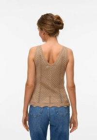 Top de punto en color beige con espalda en V, que presenta un patrón de chevron y un dobladillo con forma de concha. Combinado con jeans azules de cintura alta.