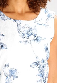 Primer plano de una persona vistiendo una camiseta sin mangas blanca con un patrón floral azul y con el cabello castaño rizado visible.