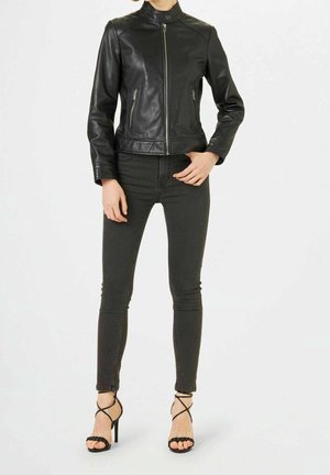 Person wearing black fitted leather jacket, dark skinny jeans, and black high-heeled sandals with ankle straps, standing on white background.