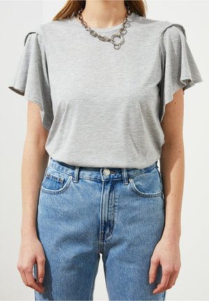 Szara koszulka z krótkim rękawem z falbankami, zestawiona z jasnoniebieskimi, wysokim stanem jeansami. Stylizację uzupełnia gruby srebrny naszyjnik łańcuchowy.