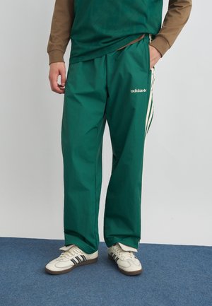 Osoba nosící zelené tepláky Adidas s bílými pruhy, bílé tenisky Adidas s černými pruhy, stojící na modrém koberci.