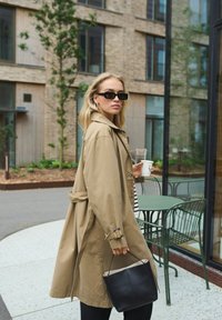 Bežový trenčkot s opaskom v páse, dlhými rukávmi a jemnou textúrou. V ruke čierna kabelka a hrnček; v pozadí zeleň a sklo.