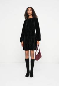 Musta sametti mekko pitkillä hihoilla, vyötäröltä kapea; yhdistetty polven ylle ulottuviin mustiin saappaisiin ja pieneen viininpunaisiin käsilaukkuun.