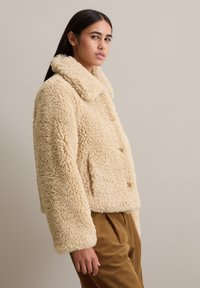 Veste en shearling crémeuse et duveteuse avec un large col, une fermeture à boutons et des poches latérales. Silhouette courte et carrée. Finition texturée et douce.