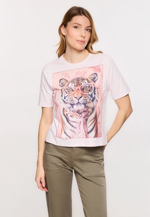 Mujer que lleva una camiseta rosa claro con un gráfico de un tigre con gafas y una bufanda estampada, combinada con pantalones verde oliva.