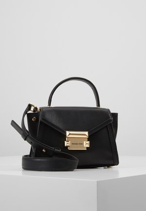 Sac à main en cuir noir Michael Kors avec fermoir doré, poignée supérieure et bandoulière réglable sur surface blanche sur fond gris.