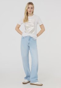 T-shirt bianca con un design a stampa leopardata lucida e testurizzata, abbinata a jeans azzurri chiari, larghi e scarpe da ginnastica bianche.