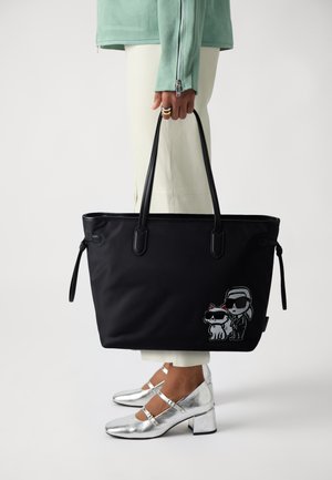 Borsa a spalla nera con due personaggi dei cartoni animati stampati di lato; tessuto morbido, manici robusti e texture liscia. Scarpe argentate visibili.