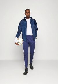 Marineblaue Sportjacke und Hose mit weißen Seitenstreifen, kombiniert mit einem weißen Hemd. Hält einen Fußball; trägt schwarze Stollenschuhe.