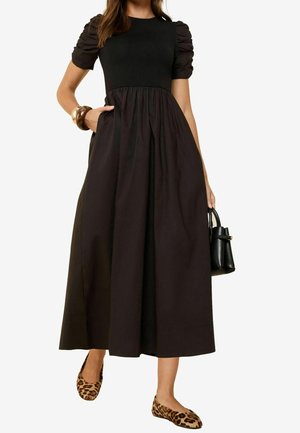 Zwarte jurk met puffmouwen, een aansluitend lijfje en een vloeiende rok; het model houdt een kleine zwarte handtas vast en draagt leopardprint ballerina's.