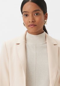 Blazer rosa chiaro con rever a toppa sopra un dolcevita a coste color crema. La modella indossa orecchini scultorei dorati, ha la pelle liscia e i lunghi capelli scuri.