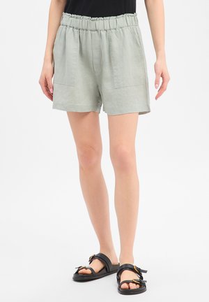 Kobieta w jasnoszortach z elastycznym pasem i czarnych sandałach na klamrę, stojąca na białym tle.