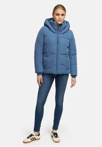 Blauwe gewatteerde jas met een grote capuchon, een voorrits, twee zijzakken en geribde boorden. Draagt met donkere jeans en crèmekleurige sneakers.