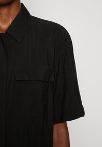 Chemise noire à manches courtes en tissu texturé, dotée d'un col pointu et d'une seule poche poitrine. Design simple et coupe décontractée.