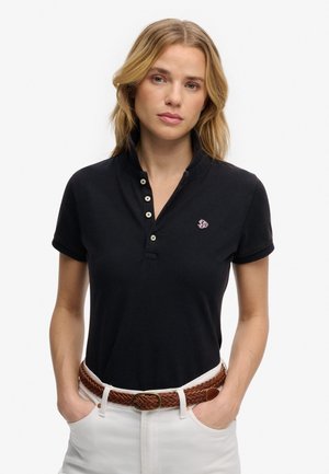 Kobieta o blond włosach, ubrana w czarną koszulkę polo z krótkim rękawem, wpuszczoną do białych spodni z brązowym plecionym paskiem, ręce w kieszeniach.