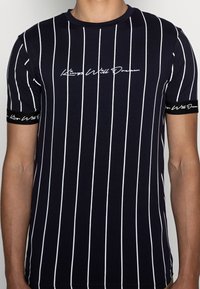 Marinblå t-shirt med kort ärm och vita vertikala ränder, med rund halsringning och svart band vid ärmarna med vit textdetalj.