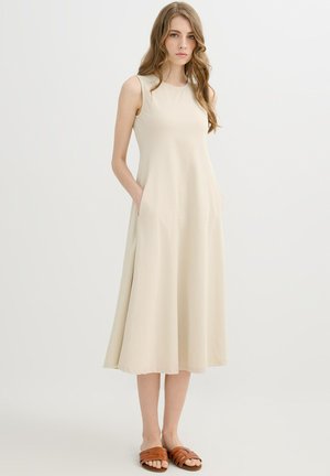 Mujer con cabello largo y ondulado, vestida con un vestido midi beige sin mangas con bolsillos y sandalias marrones con punta abierta, parada frente a un fondo blanco liso.