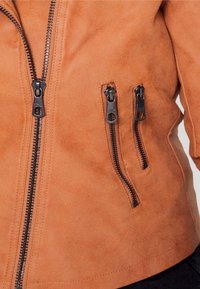Giacca in suede marrone con una texture liscia, dotata di una cerniera centrale e tre tasche frontali con cerniera, accentuate da dettagli in metallo nero.