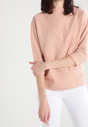 Femme portant un haut en maille côtelée pêche avec manches trois-quarts et pantalon blanc, les bras croisés sur la taille.