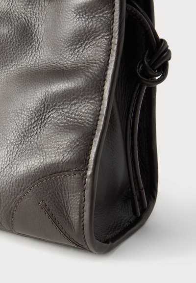 Coin d'un sac en cuir brun foncé à texture douce avec bord arrondi et détail de lanière en cuir nouée sur fond blanc.