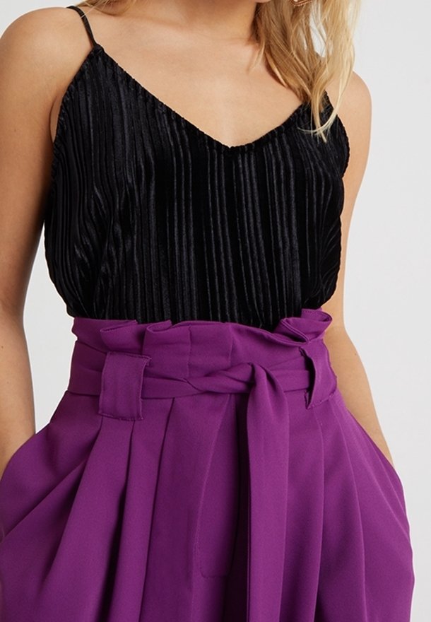 Camisole noire plissée à fines bretelles associée à un pantalon taille haute violet style paperbag avec ceinture nouée et poches latérales.