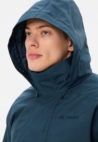 Giacca blu navy con colletto alto e cappuccio regolabile, realizzata in un tessuto morbido e impermeabile. Presenta un logo discreto.