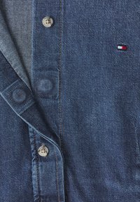 Denim shirt with a dark blue fabric, featuring a button closure and a small red, white, and blue logo. Light denim collar and stitching.