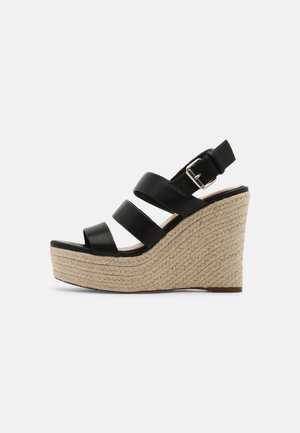 Sort kile-sandal med tre læderstropper, flettet juteplatform og spænde lukning på hvid baggrund.