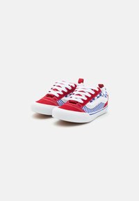 Scarpe da ginnastica in tela rossa con lacci bianchi, caratterizzate da un design a scacchi blu e bianco sul lato e una suola in gomma bianca a contrasto.