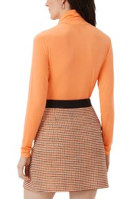 Oranje langwerpige top met hoge kraag, gecombineerd met een gemusterde mini-jurk met een gestructureerde weving in oranje en zwarte tinten.
