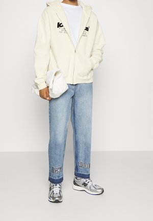 Osoba ubrana w białą bluzę z zamkiem, jasnoniebieskie dżinsy z napisem przy kostkach, srebrne trampki, trzymająca złożoną białą kurtkę.