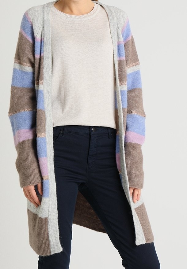 Femme portant un long cardigan rayé aux tons bleus, marron et roses par-dessus un pull beige et un pantalon foncé.