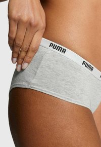 Nahaufnahme einer Hand, die auf der Hüfte ruht und helle graue Puma-Unterwäsche mit weißem Bund und schwarzem Logo-Text trägt.