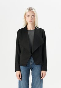 Svart, teksturert jakke med åpen front, bred krage og lange ermer, parret med en grå topp og blå jeans.