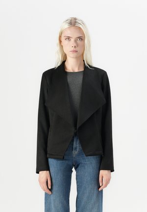 Svart, teksturert jakke med åpen front, bred krage og lange ermer, parret med en grå topp og blå jeans.