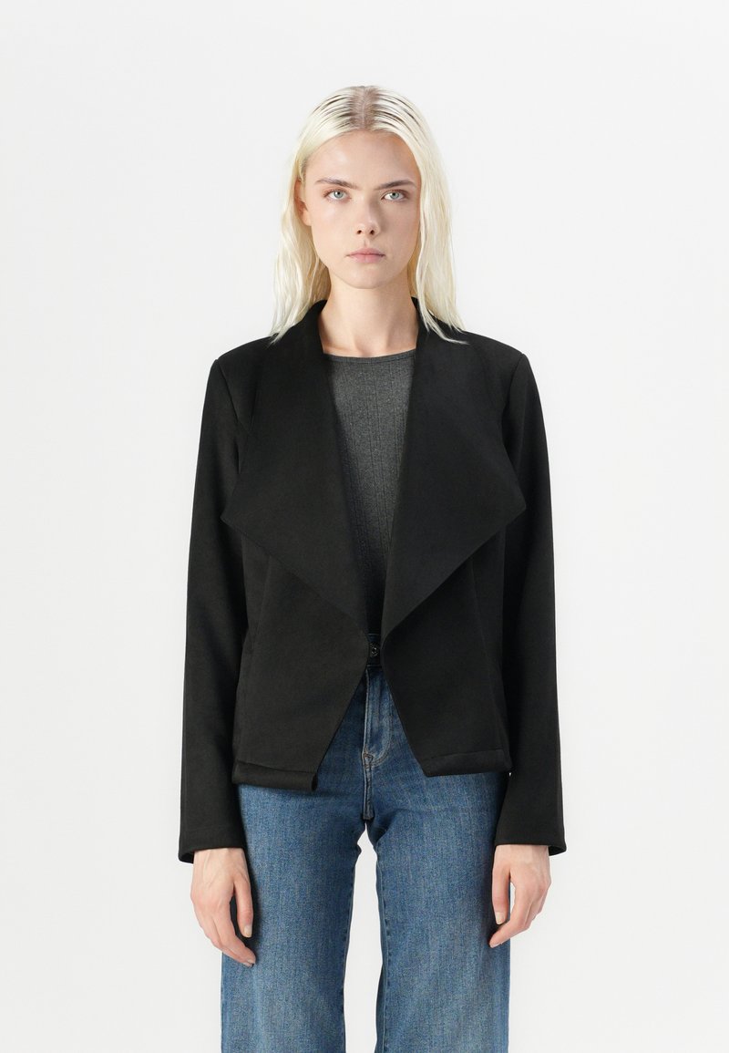 Svart, teksturert jakke med åpen front, bred krage og lange ermer, parret med en grå topp og blå jeans.