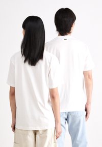 Hvit bomull t-skjorter med korte ermer, vist fra ryggen, med en liten grønn logo på nakken, kombinert med lyseblå jeans og beige shorts.