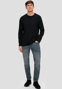 Schwarzes Langarmshirt mit einer Tasche, kombiniert mit hellblauen Jeans und schwarzen Knöchelstiefeln, auf einem neutralen grauen Hintergrund präsentiert.