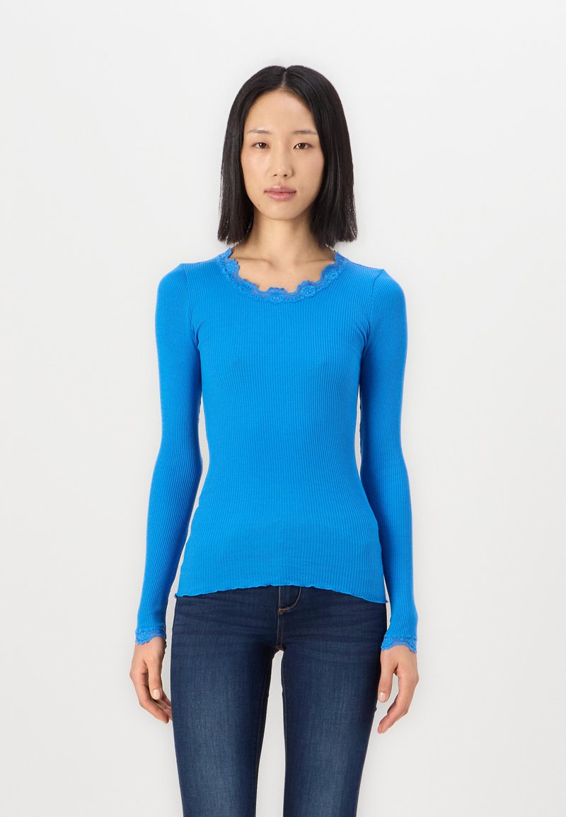 Blauw geribbelde langemouwen shirt met scalloped halslijn en manchetten. De stof lijkt zacht en aansluitend, gecombineerd met donkere spijkerbroek.