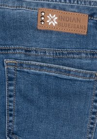 Denimjeans i medelblå nyans med en tan läderlapp som har texten "INDIAN BLUEJEANS" och tre silvernitar. Texturerat tyg.