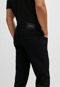 Black denim jeans with a slim fit, featuring a signature logo patch at the waistband, back pockets, and a smooth texture.