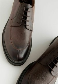 Chaussures en cuir marron avec finition texturée, bout rond, cinq œillets et semelle en caoutchouc noir. Présente des détails de couture et un design minimaliste.