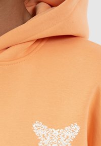 Oranje hoodie van een textuurstof. Heeft een witte bloemenvlinderprint aan de voorkant. Inclusief een verstelbare koord en een voorzak met kangoeroeontwerp.