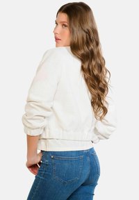 Balta teksturēta jaka ar īsu dizainu, elastīgiem manžetēm un jostasvietu, valkāta virs zilajām džinsām. Izceļ pieguļošu stilu un ikdienišķu izskatu.