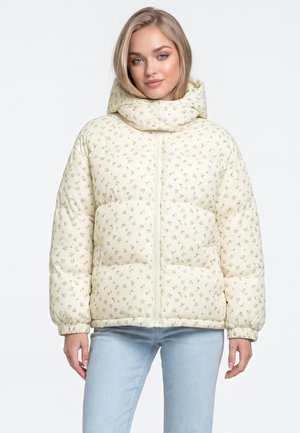 Vrouw met een lichtgele bloemen pufferjas met hoge kraag en lichtblauwe jeans, staand tegen een effen witte achtergrond.