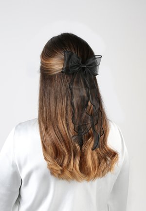 Accessorio per capelli nero trasparente a forma di fiocco con nastri arruffati, indossato su lunghi capelli castani mossi. Lo sfondo è bianco semplice.