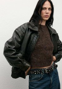 Chaqueta bombers de cuero negro con mangas oversized, suéter marrón texturizado y vaqueros azules de cintura alta con un cinturón negro con tachuelas.