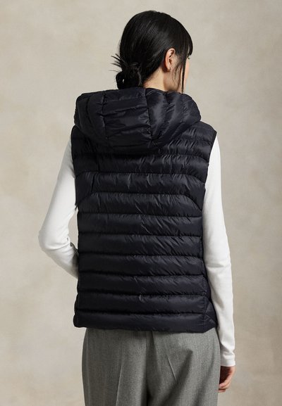 Femme vue de dos, portant un gilet noir matelassé à capuche sur un haut blanc et un pantalon gris.