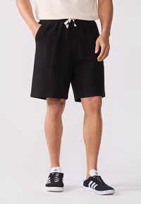 Schwarze, gerippte Shorts mit einem elastischen Bund und Kordelzug; Seitentaschen; kombiniert mit schwarzen Sneakers mit weißen Streifen.