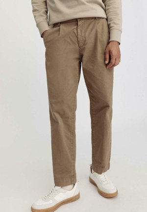 Beige Baumwollhosen mit entspannter Passform, geplooteter Vorderseite und seitlichen Taschen, kombiniert mit weißen Turnschuhen mit Gummisohlen.